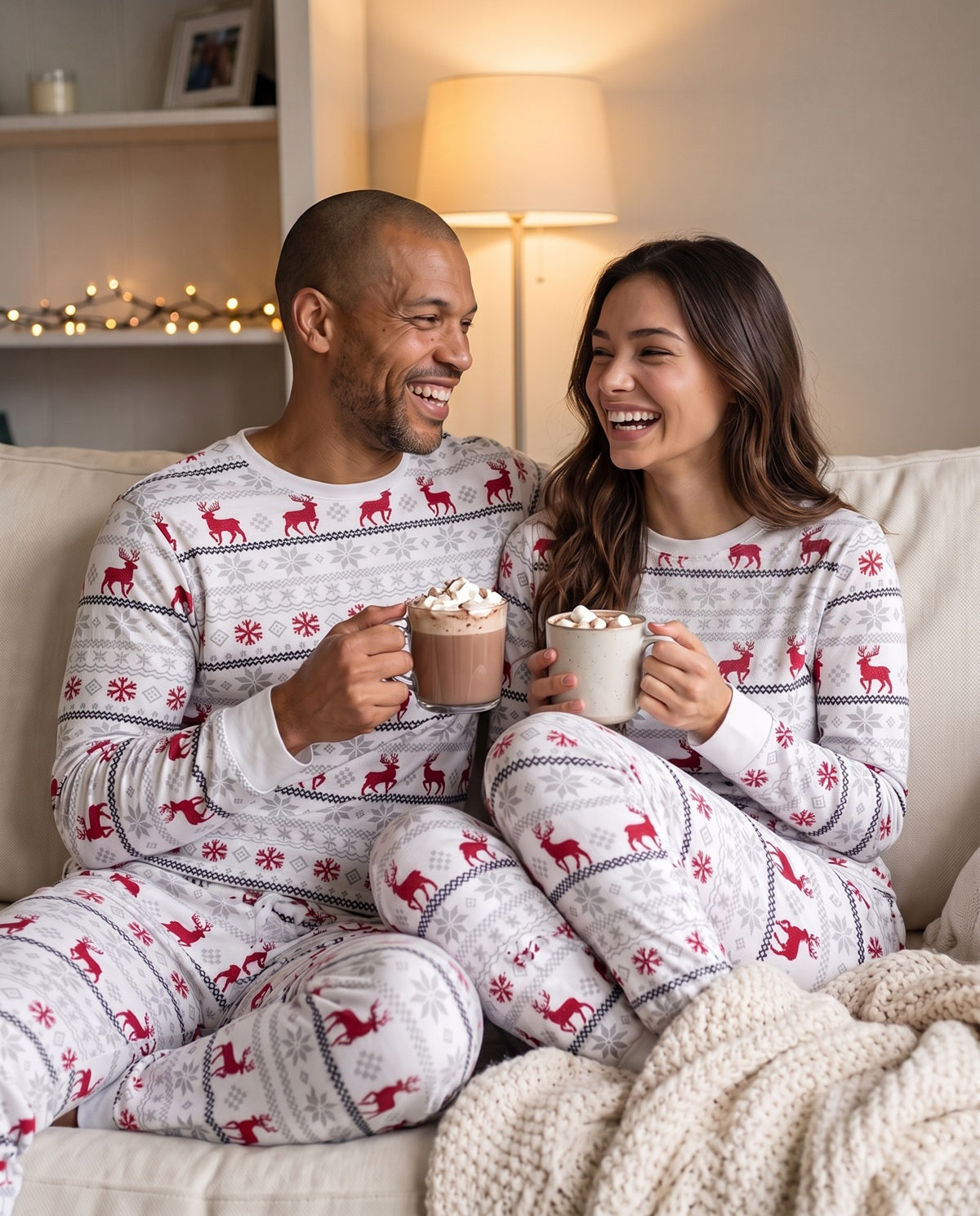 A smiling couple sitting together on a couch wearing matching white holiday pajamas with red reindeer and gray snowflake patterns, each holding a mug of hot chocolate in a cozy living room.