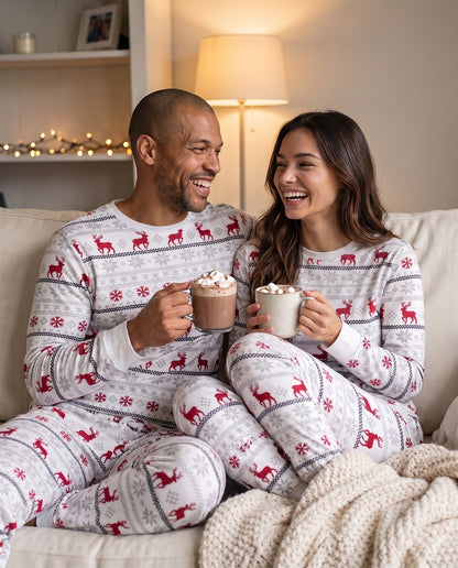 A smiling couple sitting together on a couch wearing matching white holiday pajamas with red reindeer and gray snowflake patterns, each holding a mug of hot chocolate in a cozy living room.
